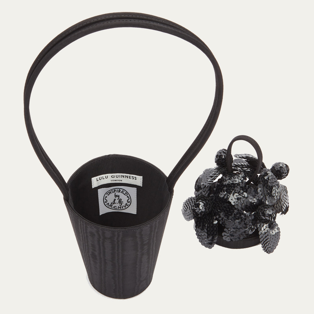 Angled side image of black satin moire bucket bag with top looped handle and black sequin flowers on the lid.