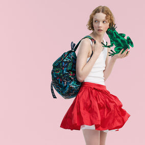 A model wearing a vibrant red pleated skirt, a white tank top, and carrying a dark blue backpack with a colorful tropical print, while holding a green frog against a solid pink background.