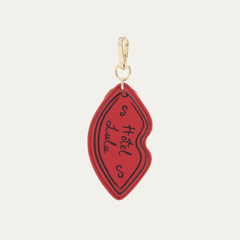 A red lip-shaped leather keyring with 'Hotel Lulu' written in white on the front, featuring shiny gold hardware.