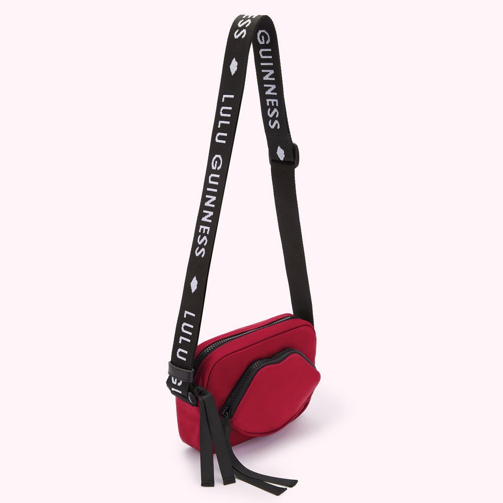 Red crossbody bag with a lip-shaped detail and black strap featuring repeating "Lulu Guinness" and "Guinness" branding.
