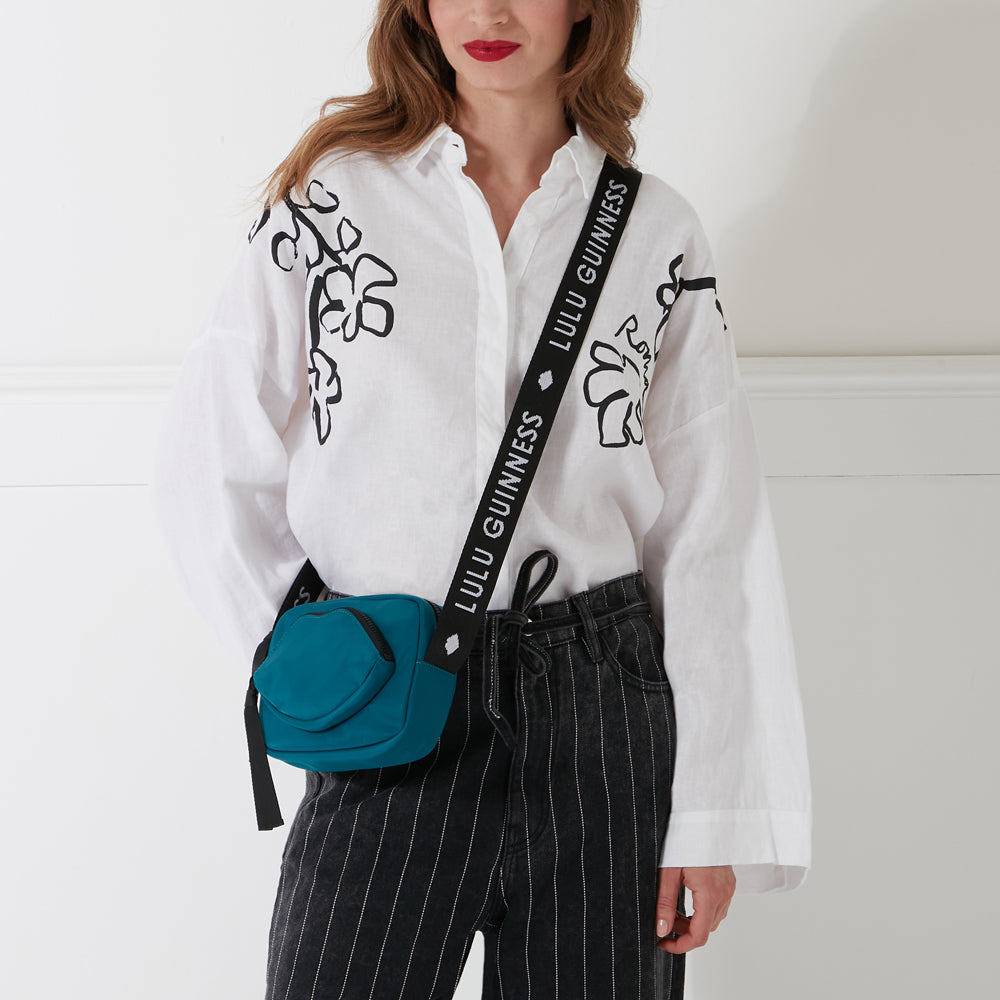 Woman with red lipstick wearing a white shirt with black floral details and dark gray pinstripe pants; she is modeling a teal crossbody bag with a black strap that says "Lulu Guinness" on it. The background is white.
