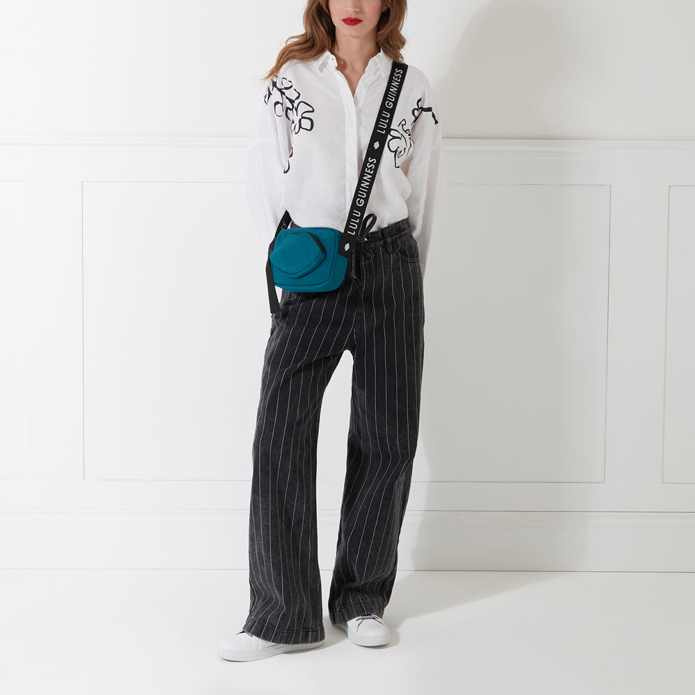 Woman wearing a white shirt, black pinstripe pants, white sneakers, and a teal crossbody bag with a black strap that says Lulu Guinness. The background is white.
