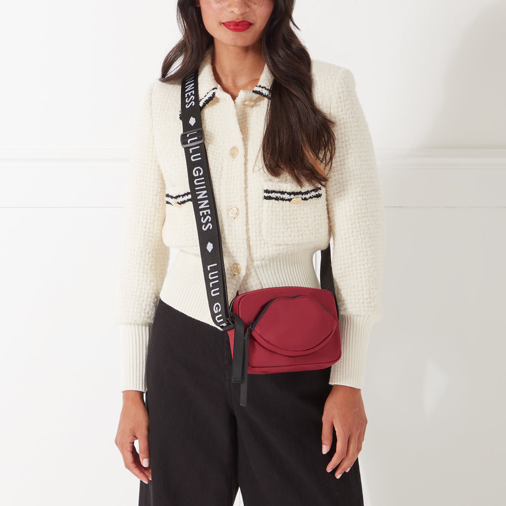 Woman wearing a cream cardigan and black pants, modeling a maroon Lulu Guinness lip-shaped crossbody bag with a black strap.
