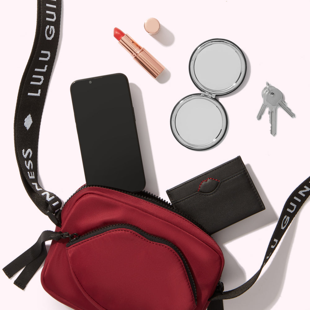 A burgundy crossbody bag with a black strap lies on a white background. Inside the bag are a black smartphone, a black cardholder, and a compact mirror. Outside the bag are a tube of lipstick and a set of keys.