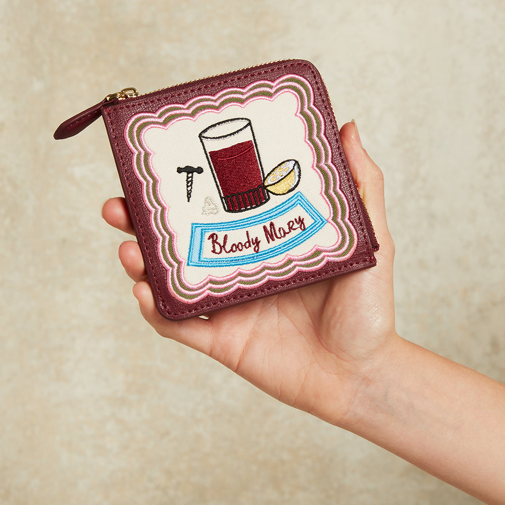 Hand holding a wallet with 'Bloody Mary' cocktail design on a beige background