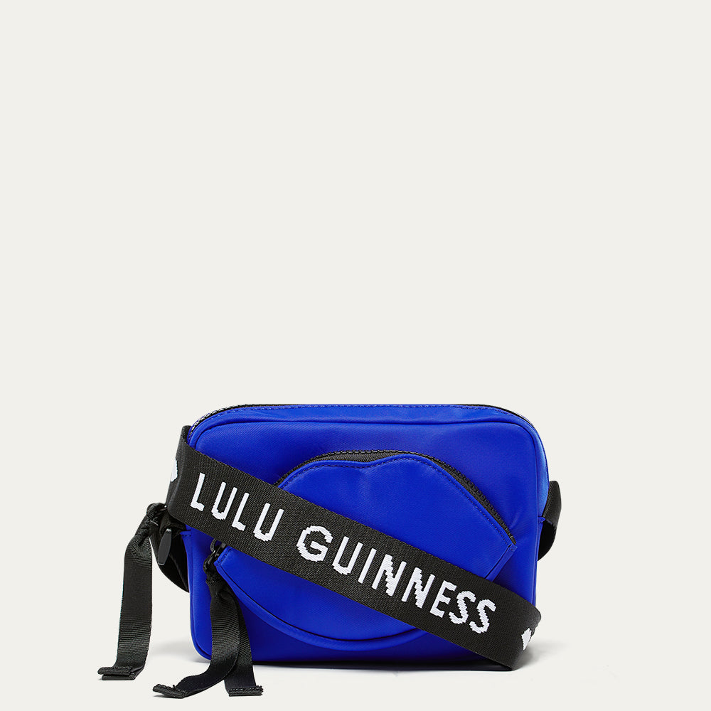 Cobalt blue nylon crossbody bag with a black branded ' Lulu Guinness' strap  and lip shaped pocket on a white background