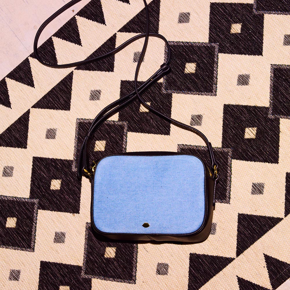 A denim and black leather crossbody bag laying on a geometric pattern rug.