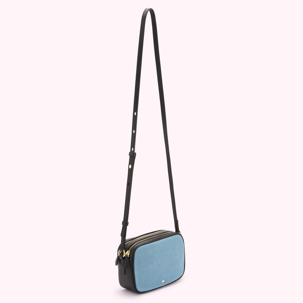 A light blue denim crossbody bag with black leather trim and a long black strap against a white background.
