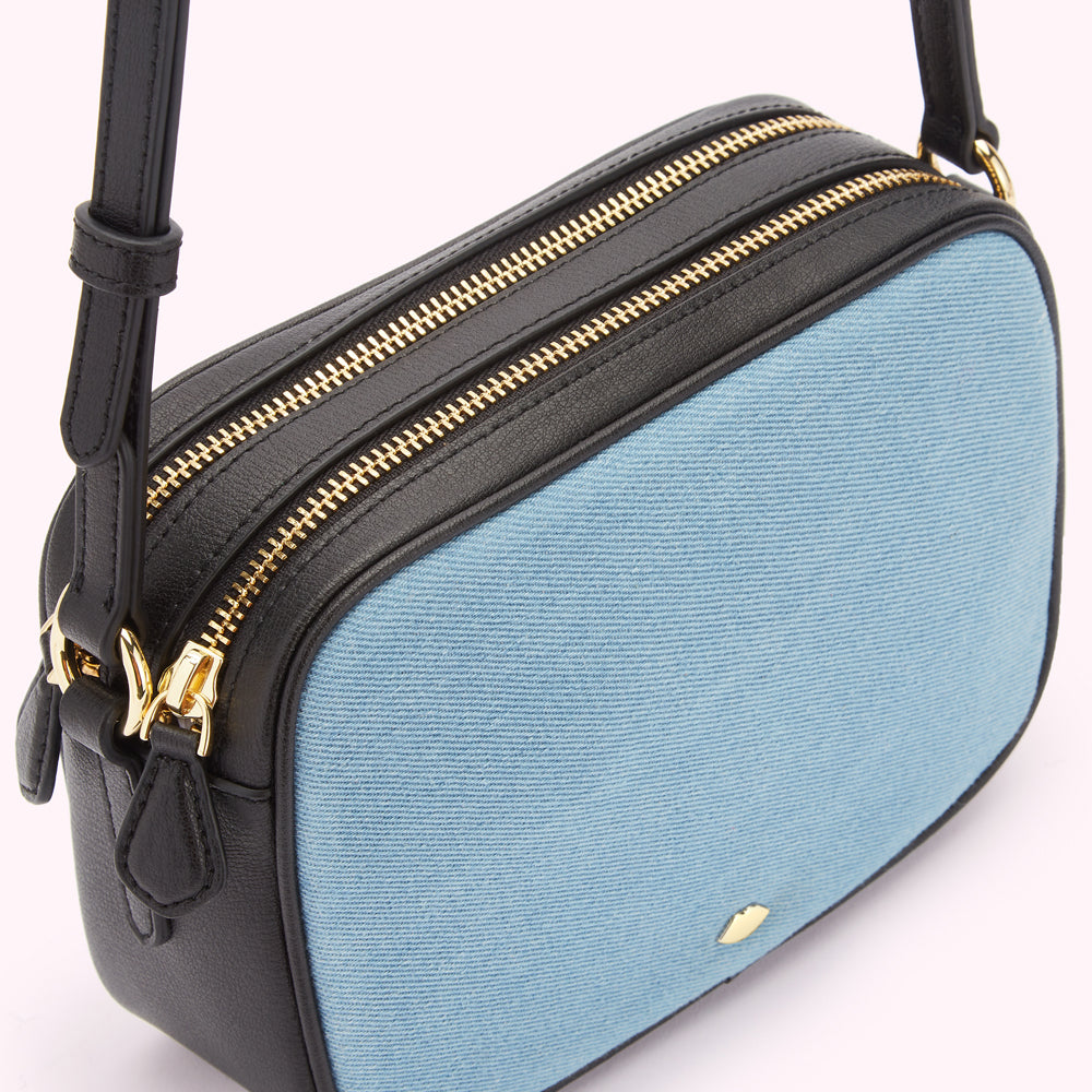 Close-up of a light blue denim crossbody bag with black leather trim and gold zippers.
