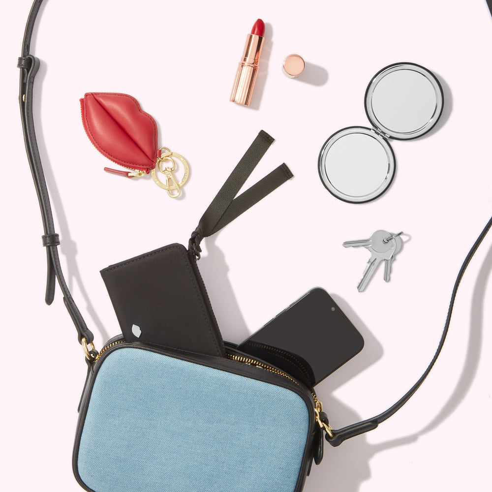 A light blue denim crossbody bag with a black cardholder, smartphone, lip-shaped keychain, compact mirror, lipstick and keys spilling out of it against a white background.
