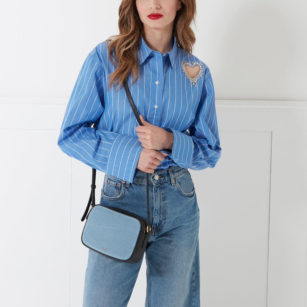 Woman in blue striped shirt and jeans with light blue denim crossbody bag and jeweled heart brooch.
