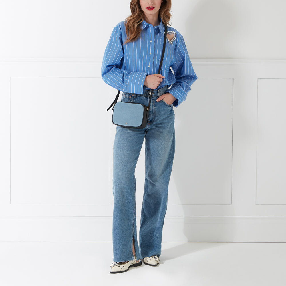 A woman in blue jeans and a blue striped shirt wears a light blue crossbody bag against a white background.