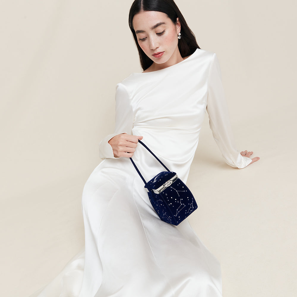 Woman in a white dress holding a blue velvet handbag featuring stars and constellations against a beige background
