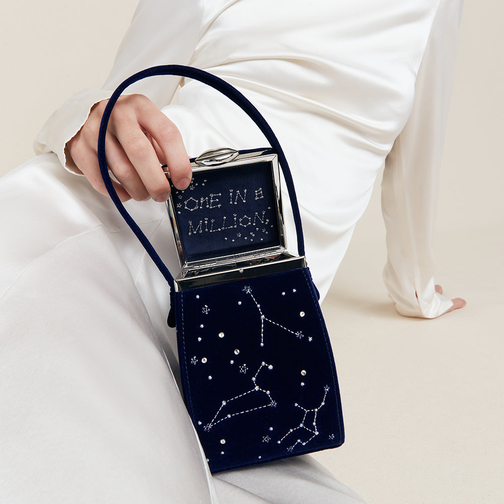 Navy blue velvet handbag with star constellations and 'One in a Million' text held by a person in white.