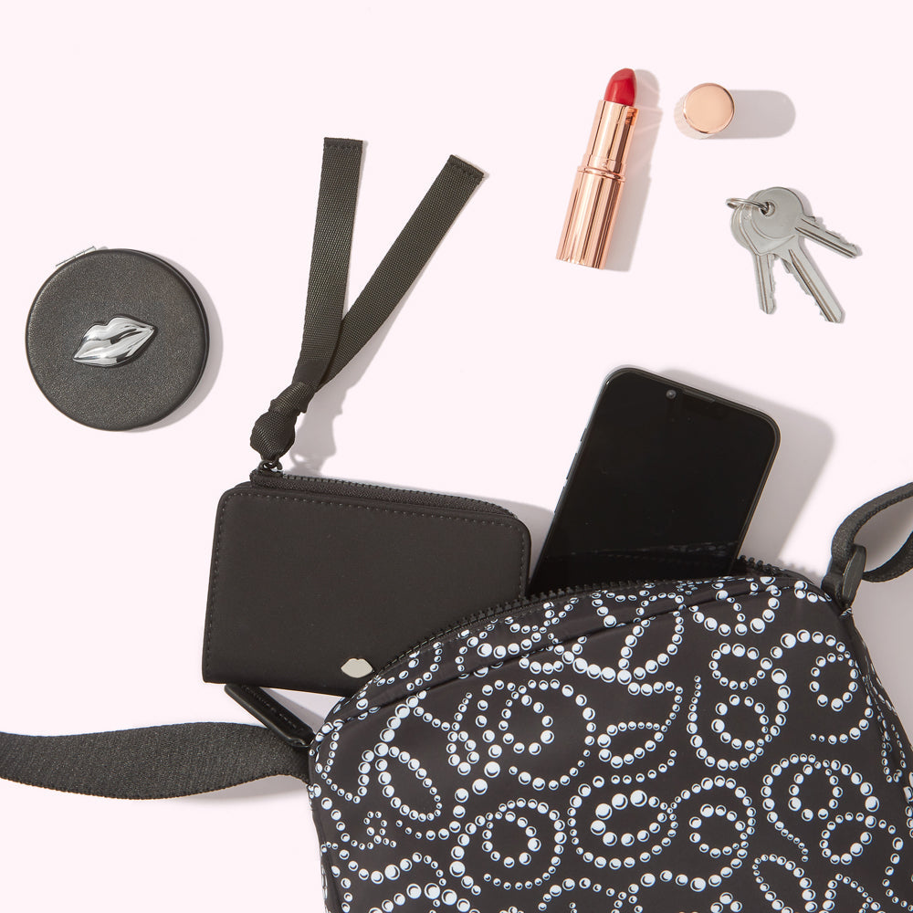 A black crossbody bag with a pearl pattern, containing a black wallet, smartphone, and a compact mirror; a lipstick and keys are next to the bag on a white background.
