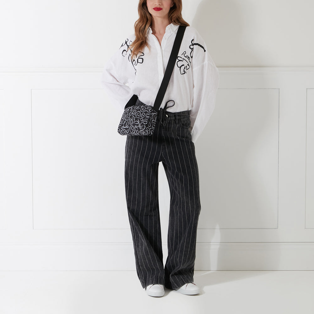 Woman wearing a white oversized shirt, black pinstripe pants, white sneakers, and a black crossbody bag with a white pattern against a white wall.
