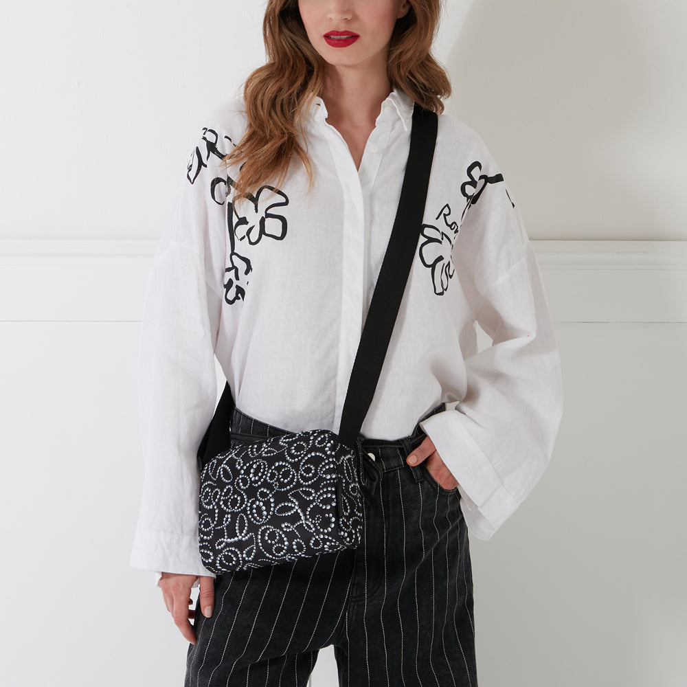 Woman wearing a white shirt with black floral print and black pinstripe pants, carrying a black crossbody bag with white embellishments. The background is white.
