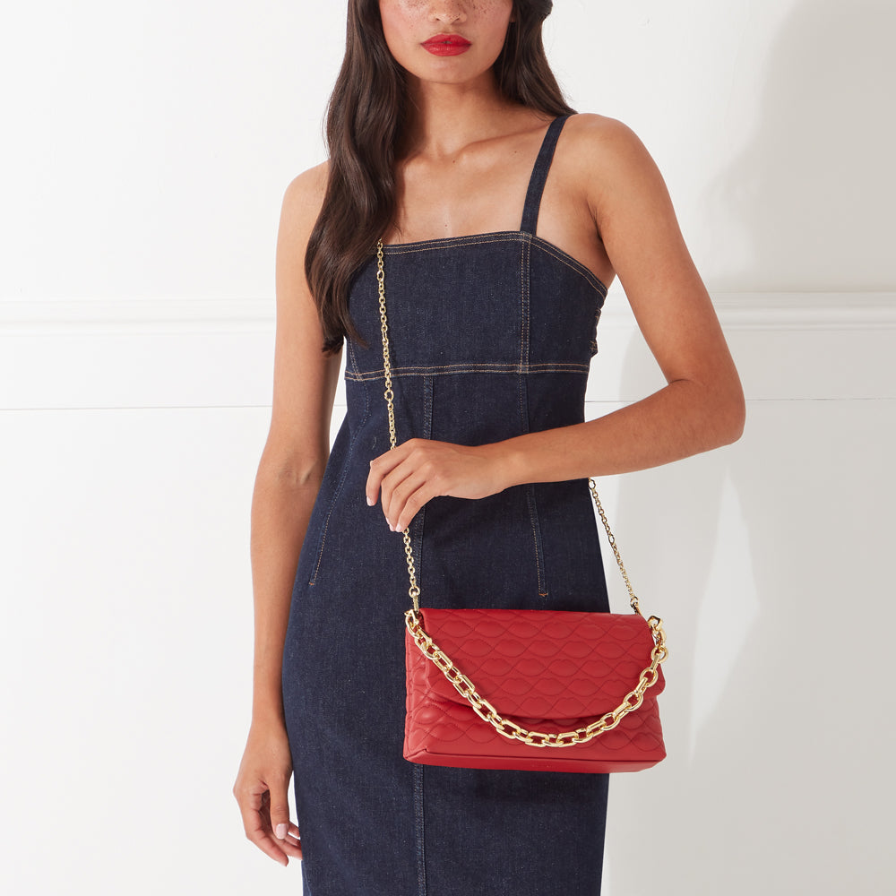 Woman in denim dress carrying red quilted crossbody bag with gold chain strap. White background.
