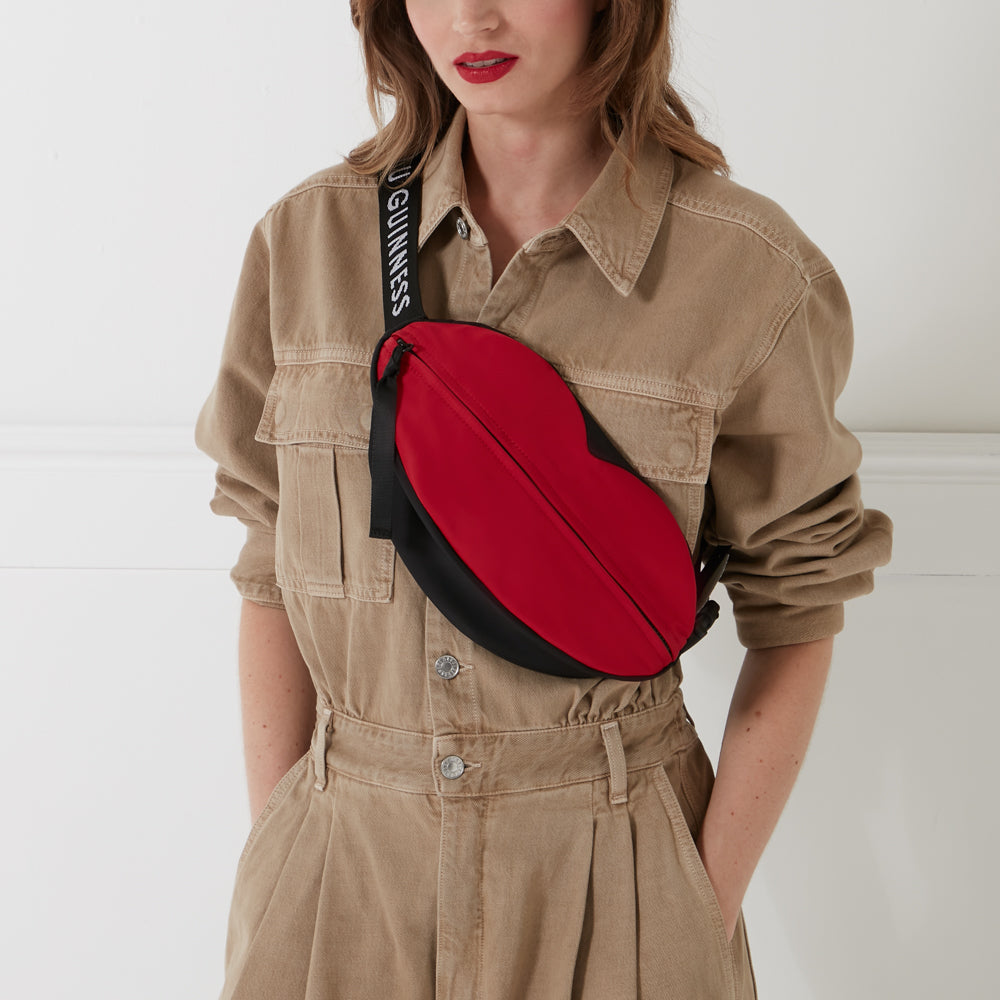 A woman in a tan jumpsuit wears a red and black lip-shaped fanny pack with a Guinness-branded strap.