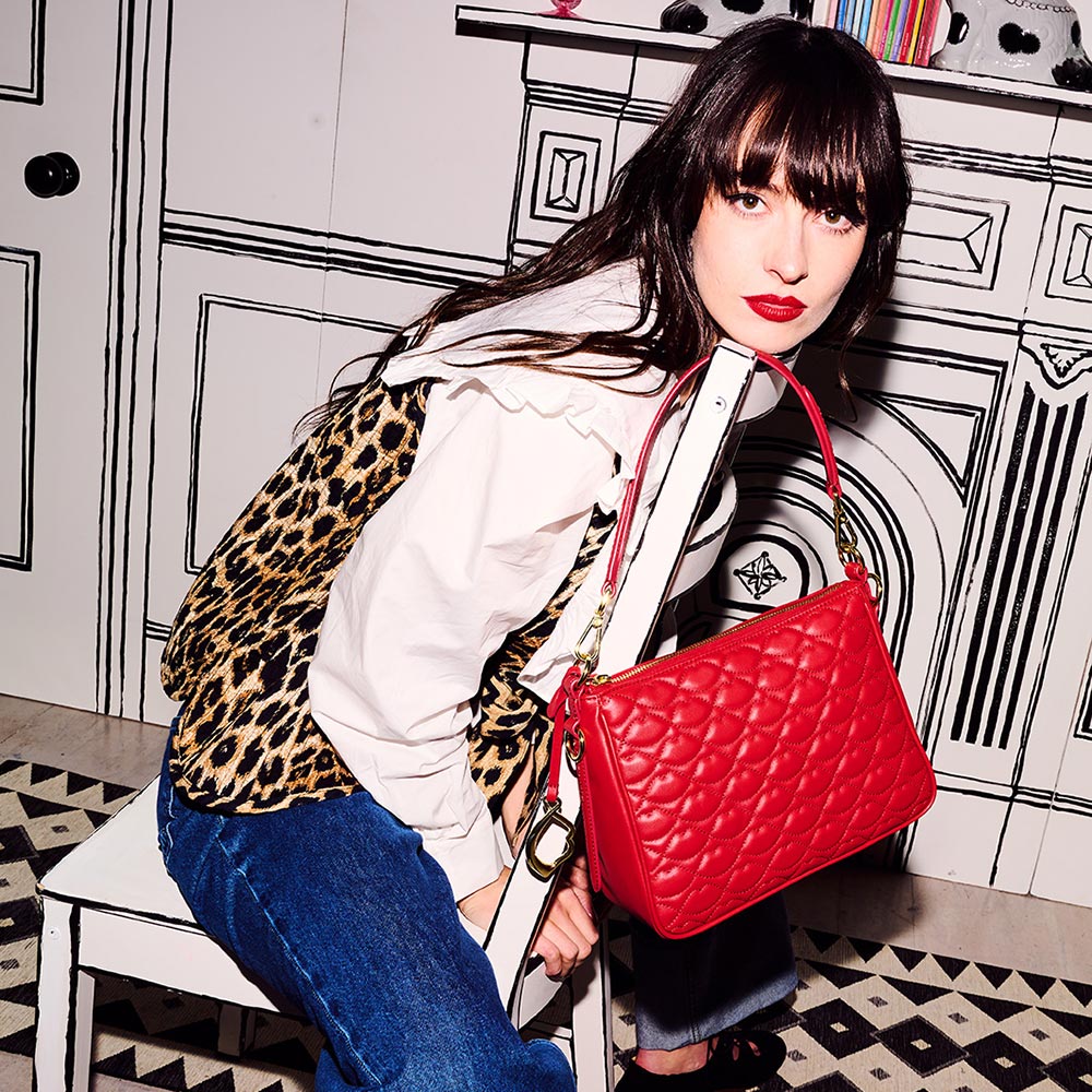 Model with dark hair sitting on a black and white chair wearing a leopard print waistcoat over a white shirt and denim jeans. A red quilted bag hangs off the chair with a lip shaped metal charm and lip shaped quilting.