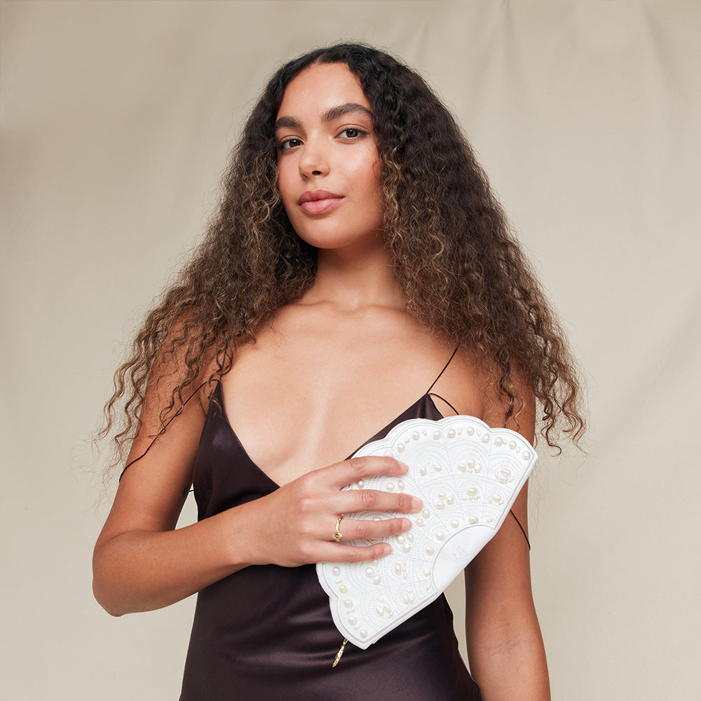 Woman holding a white fan shaped clutch with pearl embellishments against a beige background