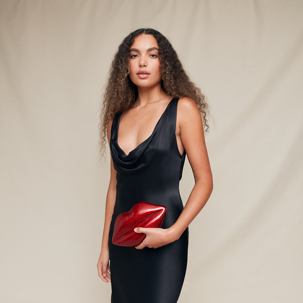 Woman in a black dress holding a red lip-shaped clutch against a beige background