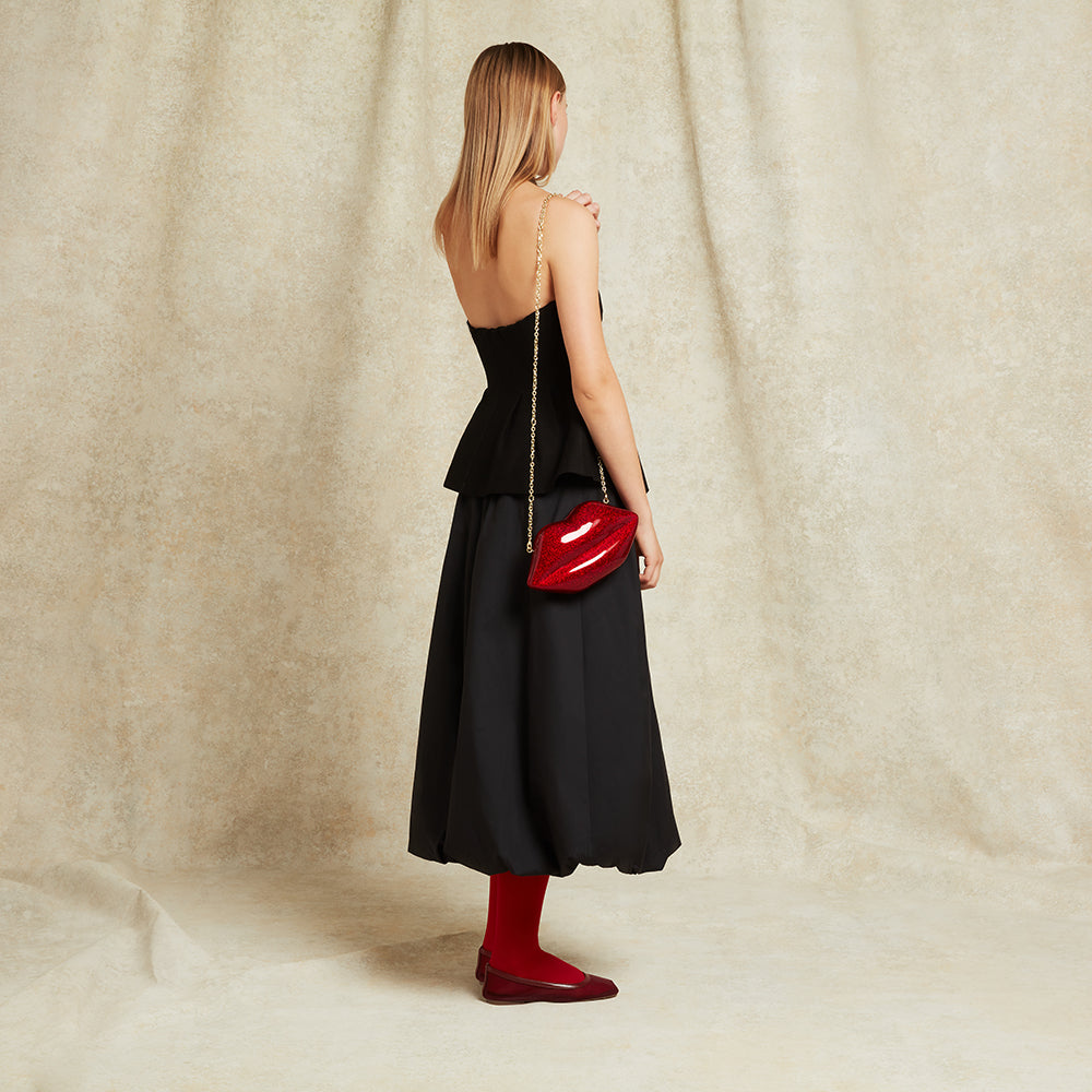 Woman in a black dress holding a red glittery lip-shaped handbag against a beige curtain background
