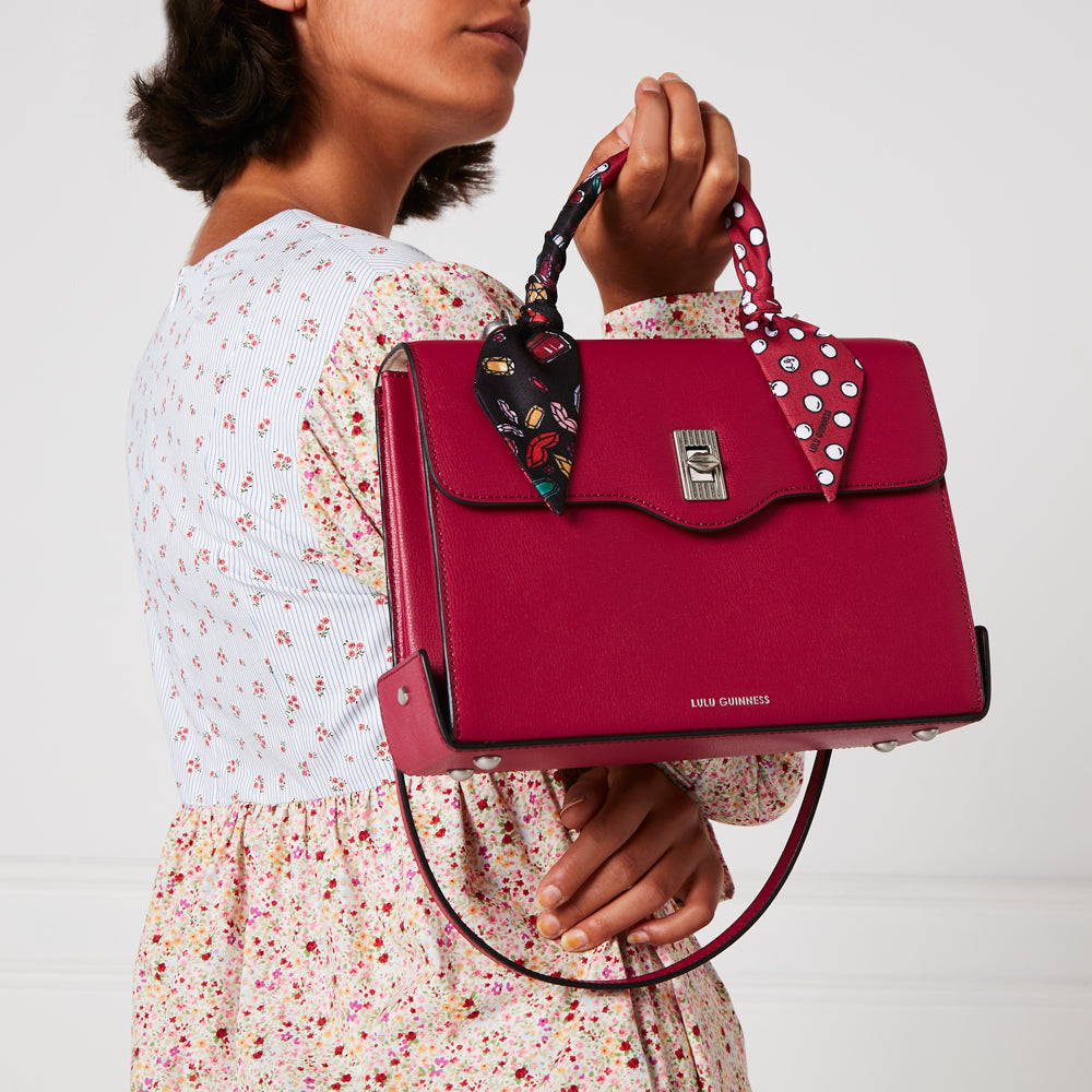 A woman in a floral dress holds a crimson Lulu Guinness satchel adorned with two patterned scarves.
