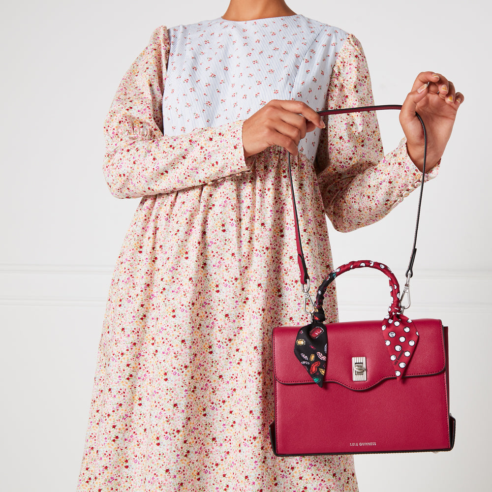 A woman in a floral dress holds a red Lulu Guinness handbag with a patterned scarf around the handle.