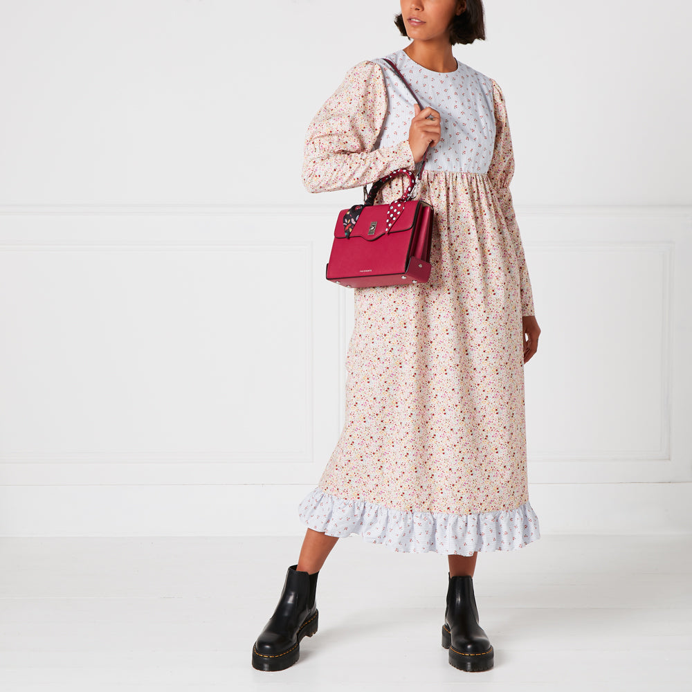 Woman in floral dress and black boots carrying a red handbag against a white background.
