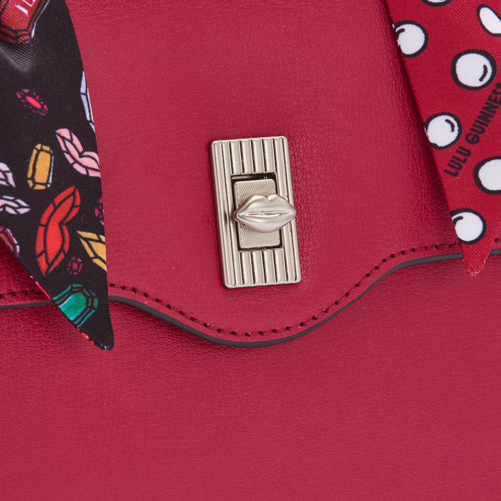 Close-up of a crimson Lulu Guinness handbag's clasp, detailed with a silver lip-shaped embellishment and patterned scarf accents.