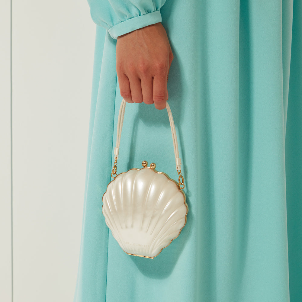 A woman in a teal dress holds a small, white seashell-shaped purse with a gold clasp.
