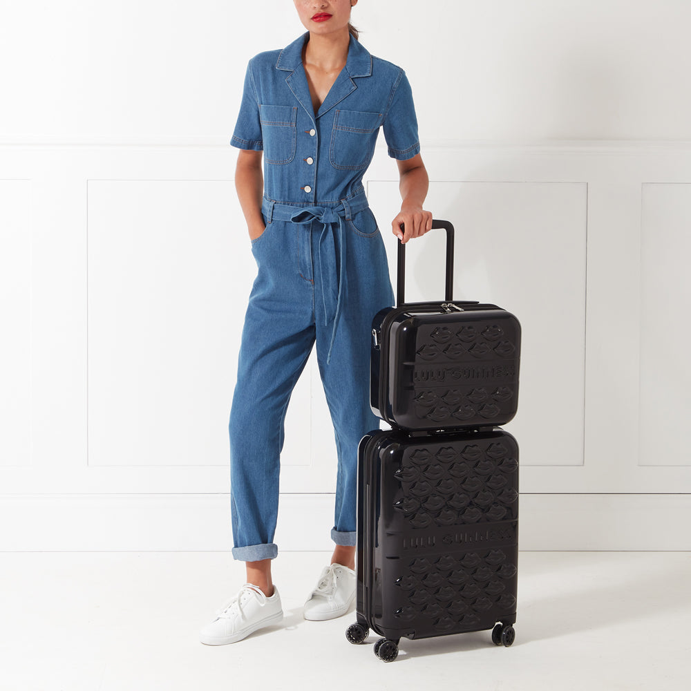 A woman in a denim jumpsuit stands next to a black two-piece luggage set decorated with a lip print.