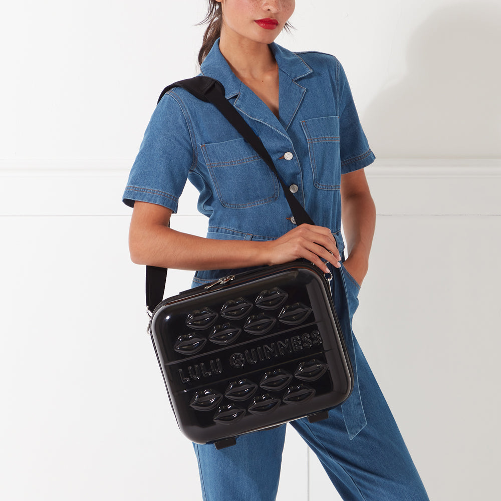 Woman in denim overalls holding a black Lulu Guinness carry-on suitcase with lips design.
