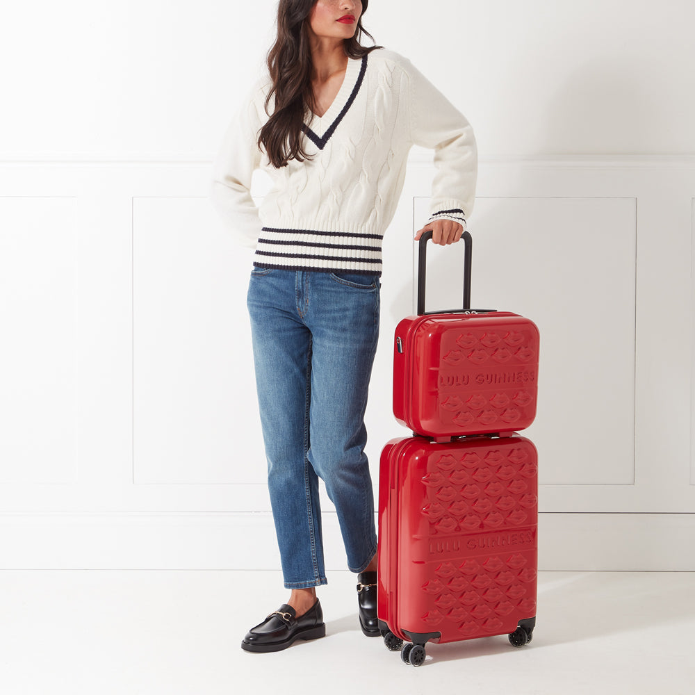 A woman in jeans and a cream sweater stands next to a red two-piece Lulu Guinness suitcase set against a white background.
