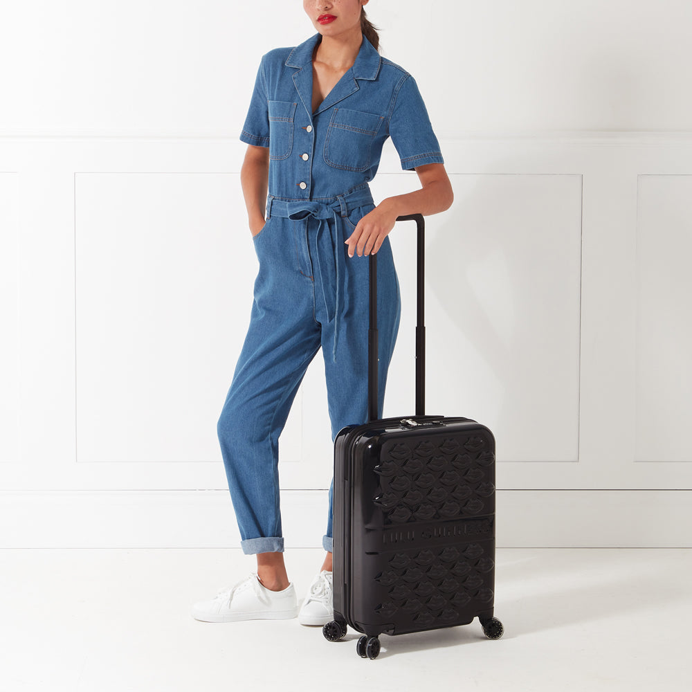 A woman in a denim jumpsuit stands next to a black Lulu Guinness suitcase with a lip print design against a white background.
