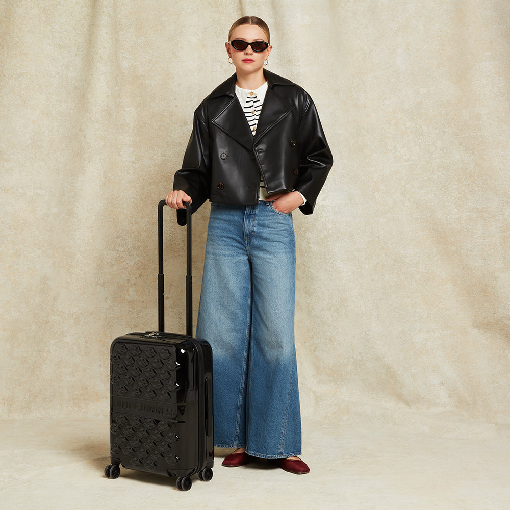 Person wearing a black leather jacket and blue jeans, holding a black suitcase against a beige background