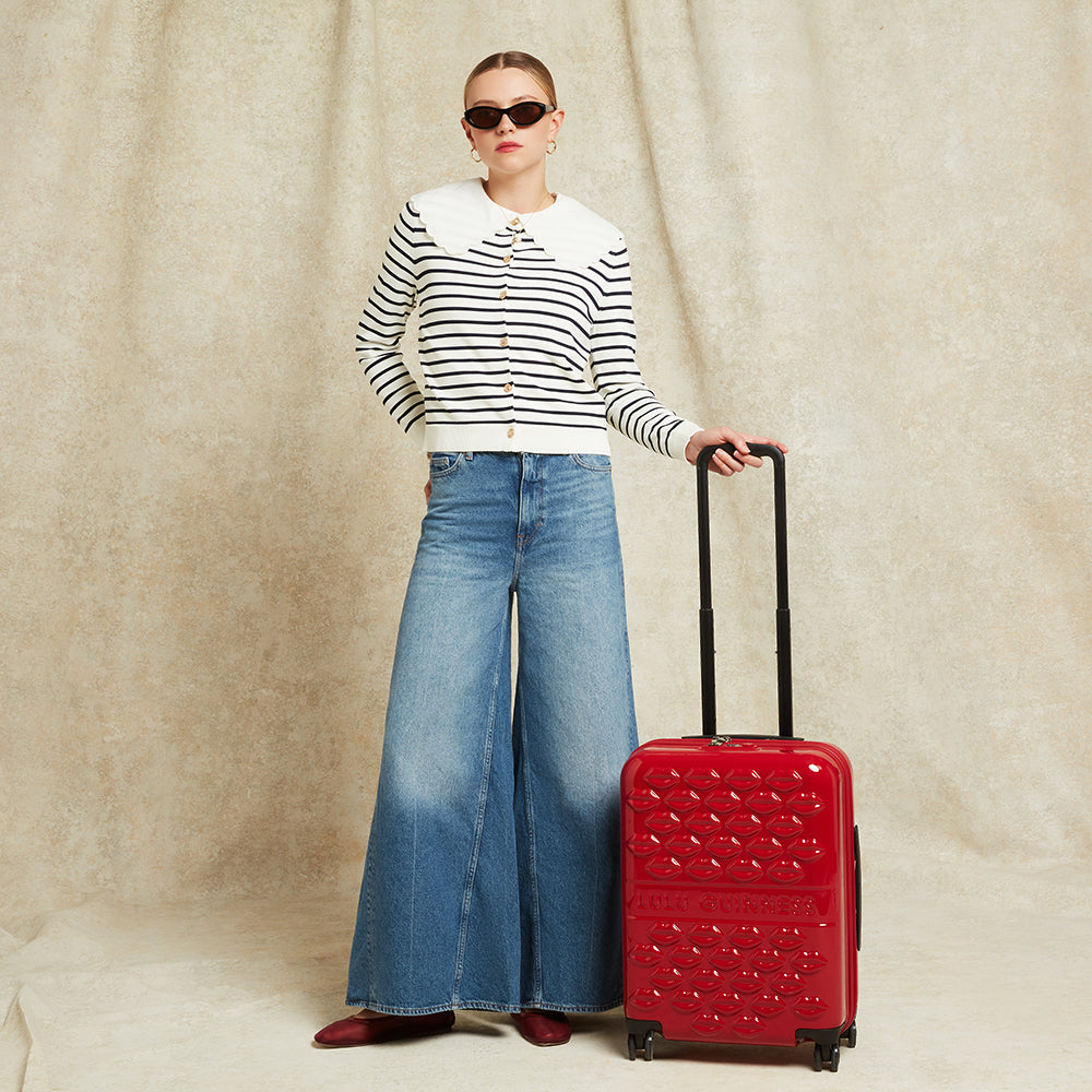 Woman in striped sweater and wide-leg jeans holding a red suitcase against a beige background