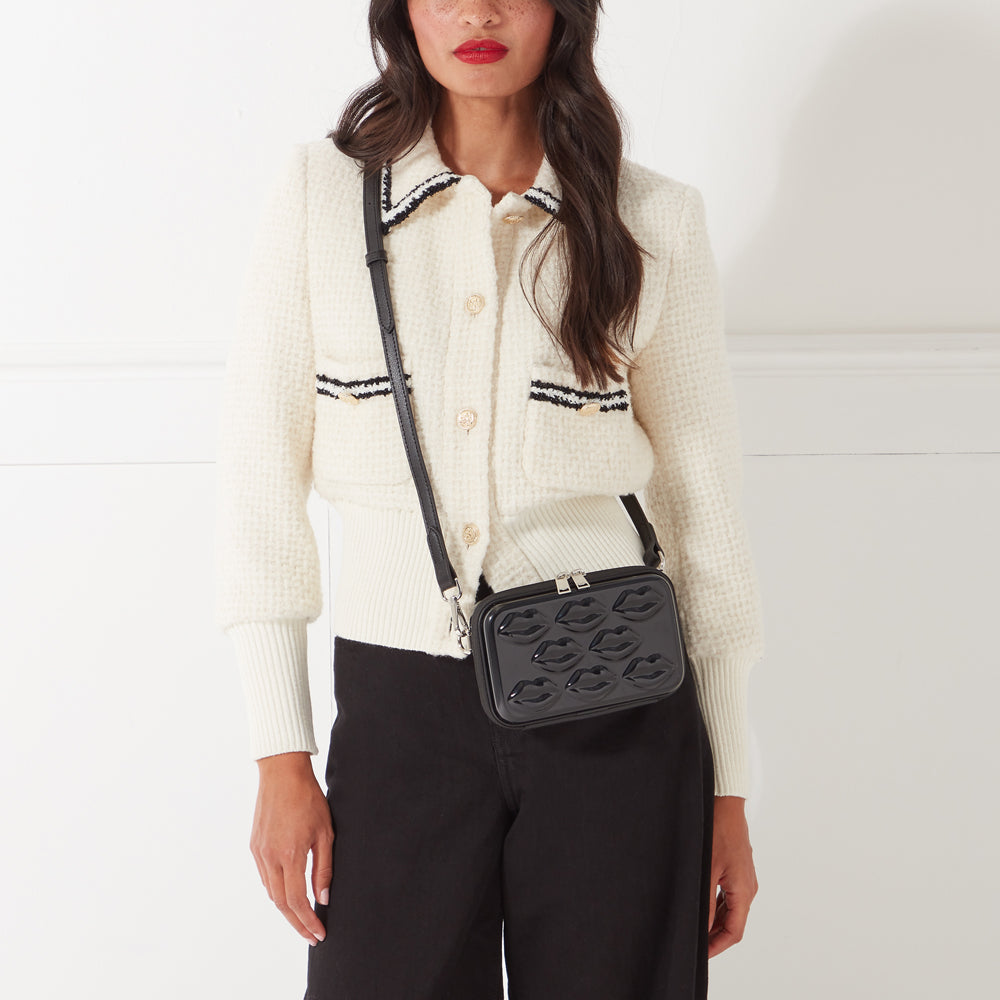 Woman wearing a cream cardigan and black pants, showcasing a black crossbody bag with lip-shaped embossing.