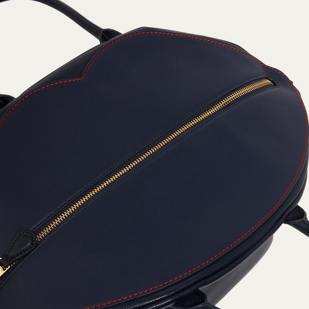 Close-up image of a navy blue leather tote bag with gold zipper and contrast red stitching on a white background