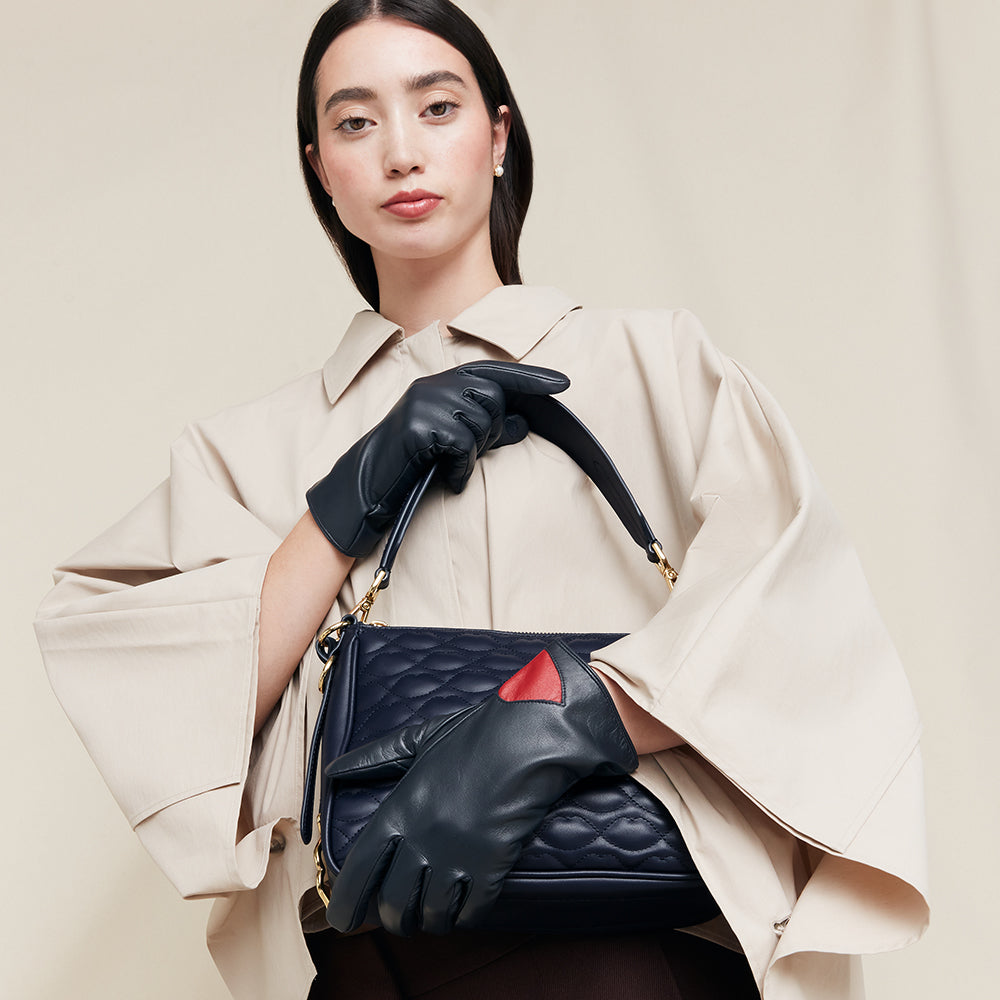 Woman holding a navy leather handbag wearing navy leather gloves with red accents, wearing a beige coat on a beige background