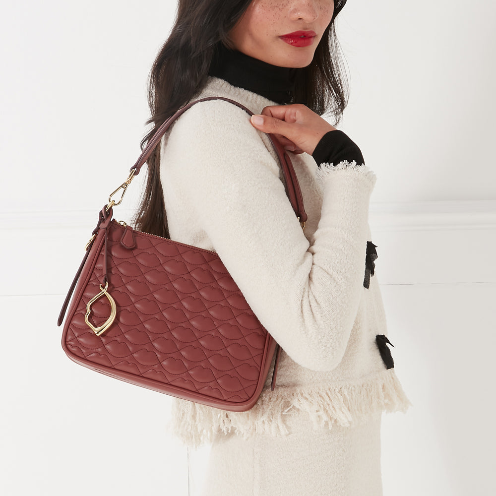 Model wearing a cream textured jacket with fringe details and holding a quilted russet shoulder bag featuring a gold lip-shaped charm, standing against a white background