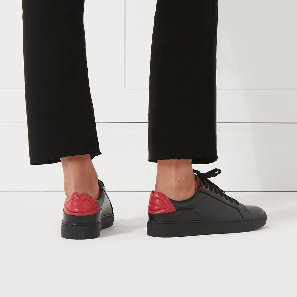 Black sneakers with red lip-shaped detail on the back, worn by a model against a white background.
