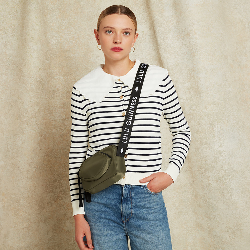 Woman wearing a striped shirt with a green bag with 'Lulu Guinness' on the strap, standing against a beige background