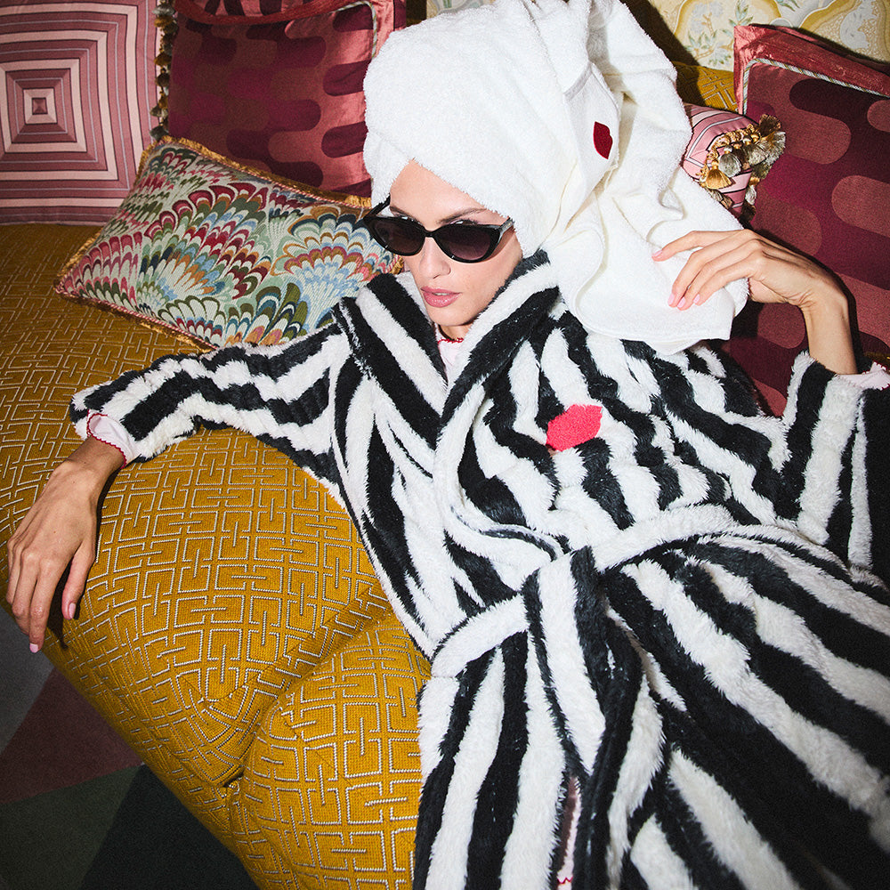 Person wearing a black and white striped robe with a red embroidered lip design, sitting on a patterned chair.
