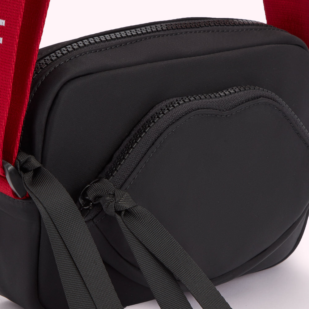 Close-up of a black crossbody bag with a red strap and a lip-shaped zipper detail.