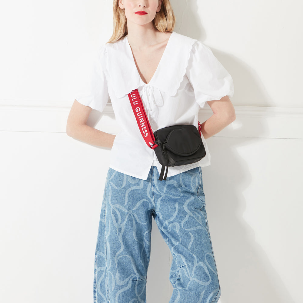 Woman with blonde hair wearing a white shirt, denim jeans and a black nylon crossbody bag with lip pocket on the front with a red 'Lulu Guinness' logo strap.