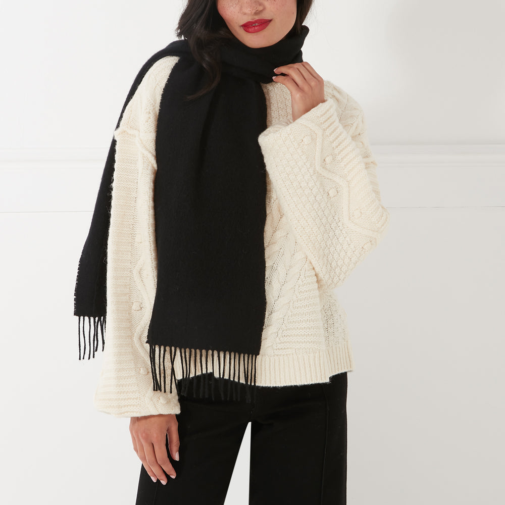 Woman wearing a black fringed scarf over a cream-colored sweater.