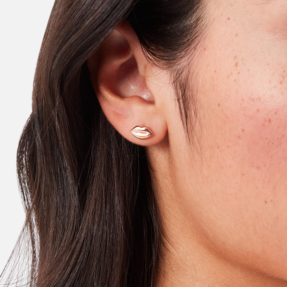 Close-up of a person's ear wearing rose gold lip-shaped stud earrings against a white background.
