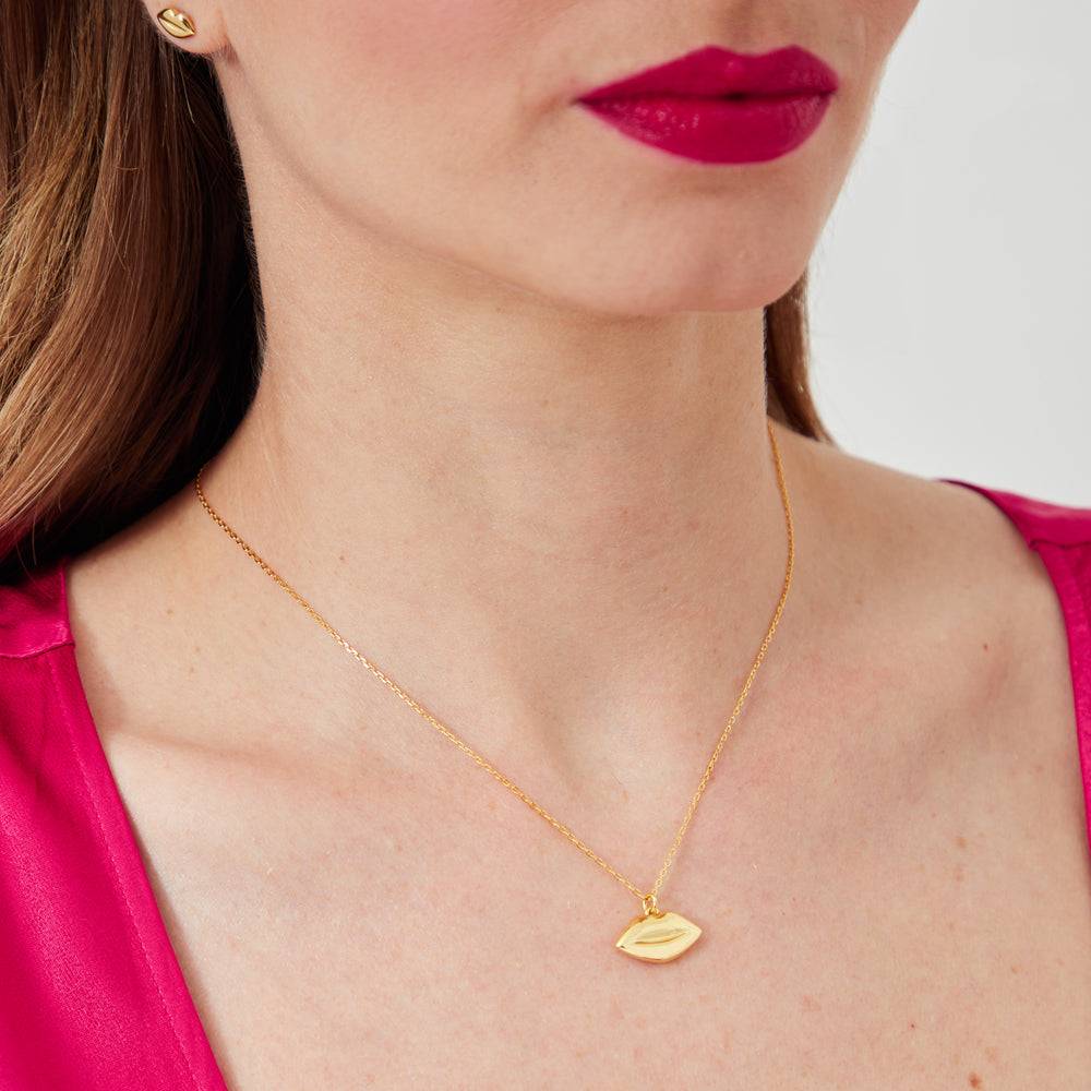 Close-up of a woman wearing a gold lip-shaped necklace and earrings against a white background. She is wearing a fuchsia top.
